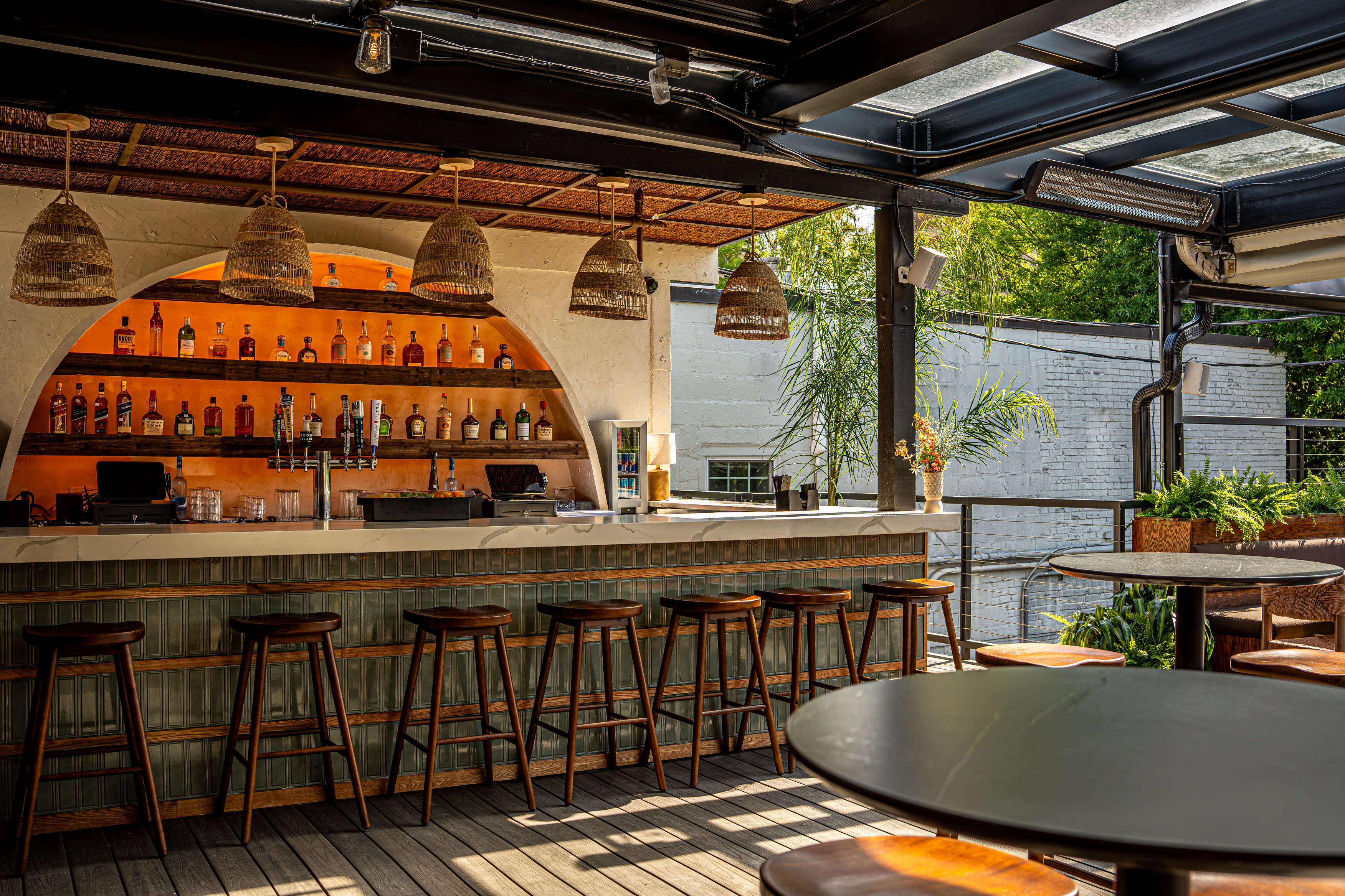 A stylish bar area featuring a white countertop, wooden stools, and various bottles displayed on shelves. The bar is illuminated with warm lighting and surrounded by greenery, creating a modern and inviting atmosphere.