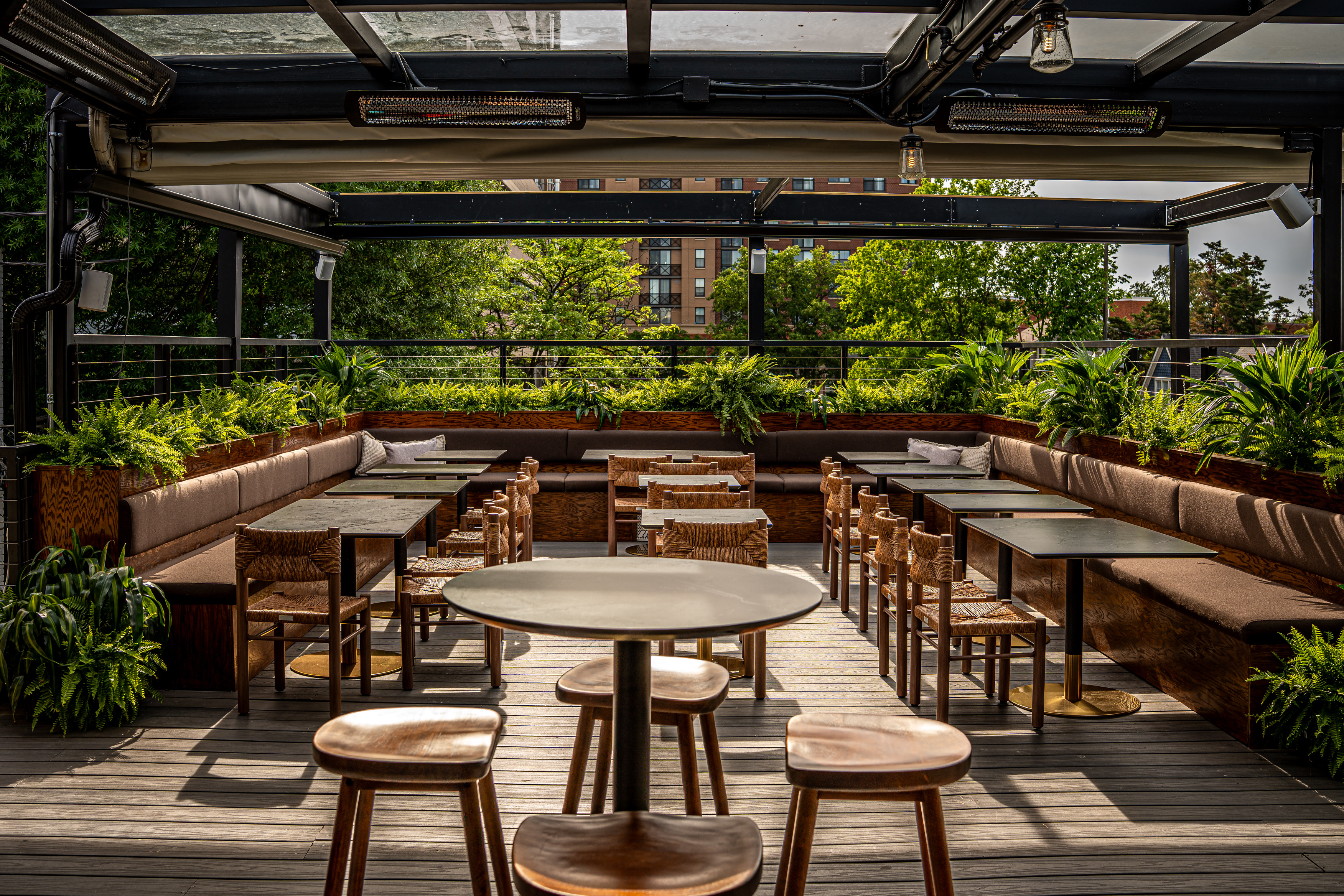 A rooftop restaurant with wooden tables and chairs, surrounded by greenery, featuring a covered seating area and warm lighting.