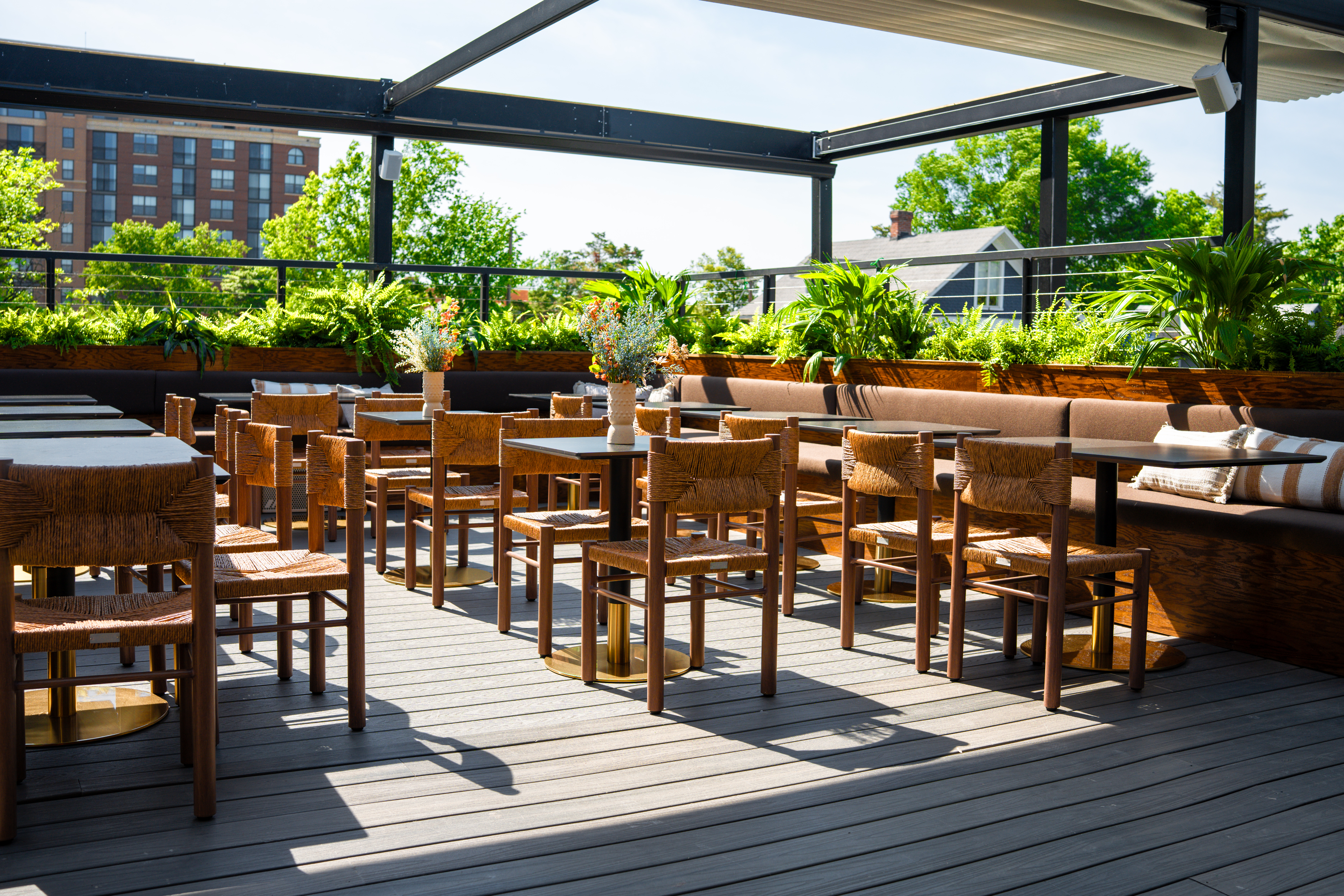Sunny rooftop dining area with wooden chairs and tables, surrounded by lush greenery and decorative plants.